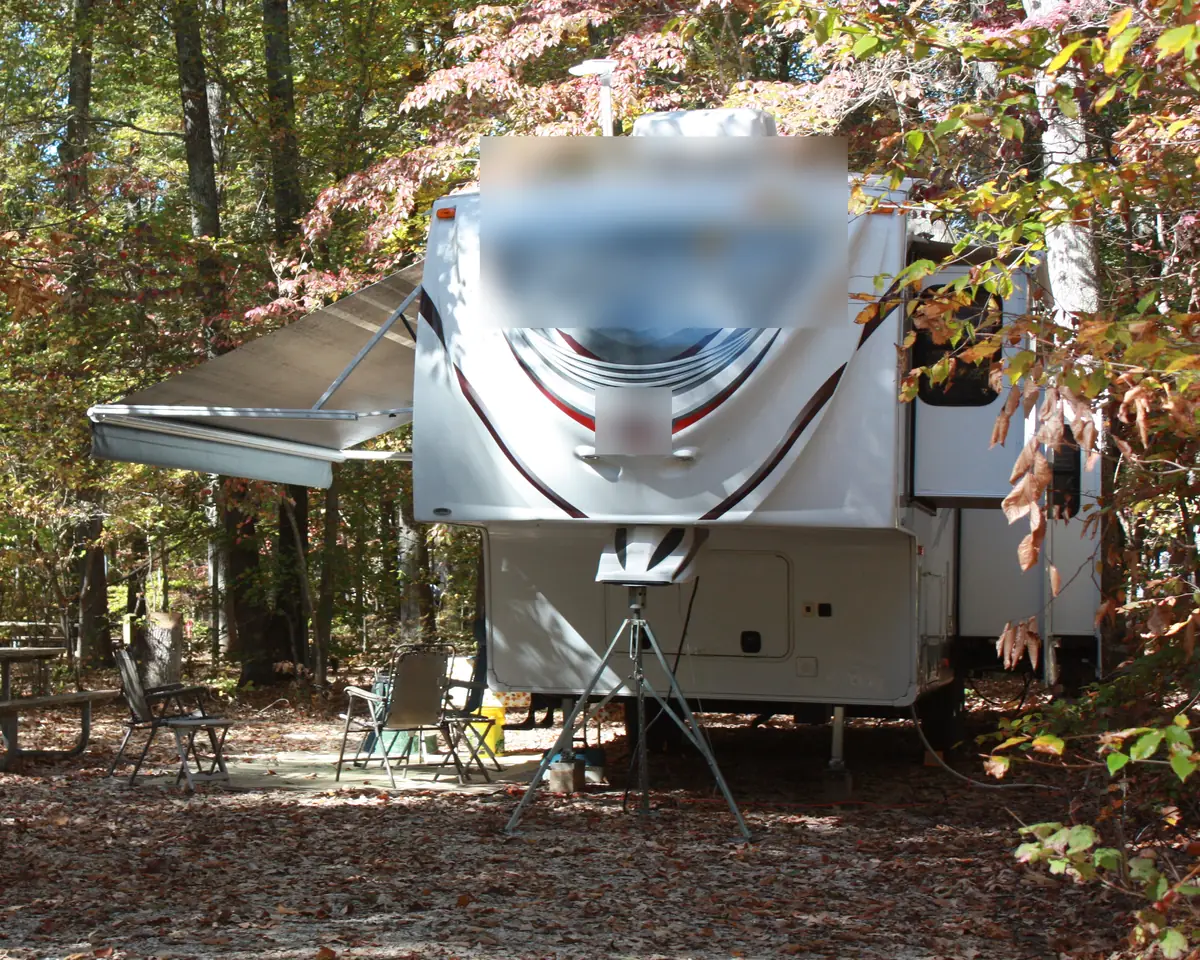Fifth wheel trailer parked at a wooded campsite with its patio awning extended and a picnic table beside it