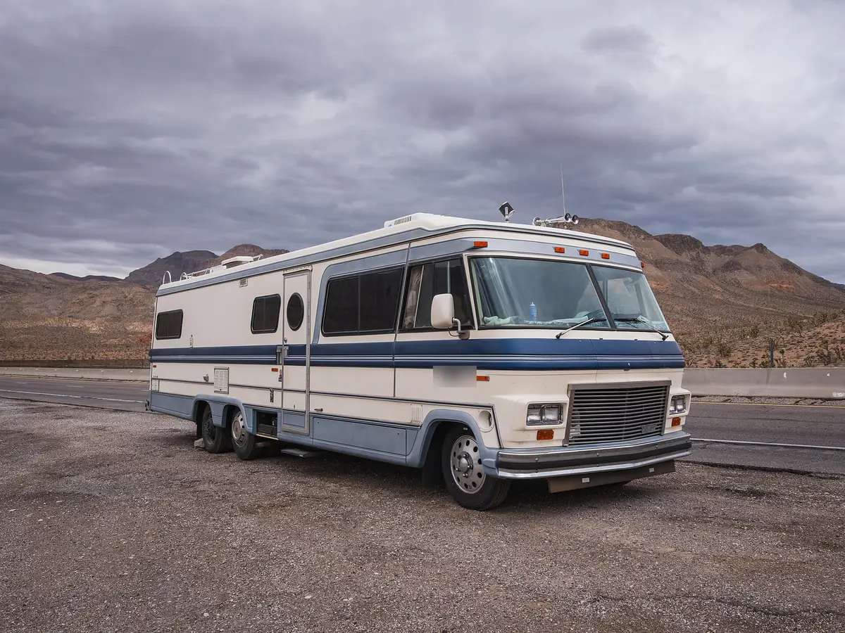 Older Class A motorhome parked on gravel beside a desert highway under gray clouds